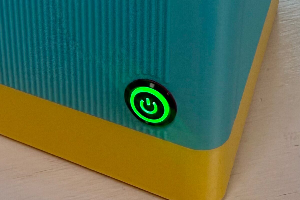 Close-up of the button on a StickyFax — a round ring glowing green against the ribbed teal shell, above the yellow base