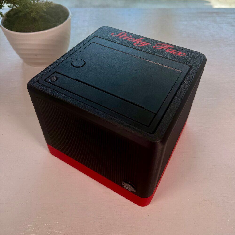 An ink-black-and-signal-red StickyFax on a desk, its power button lit