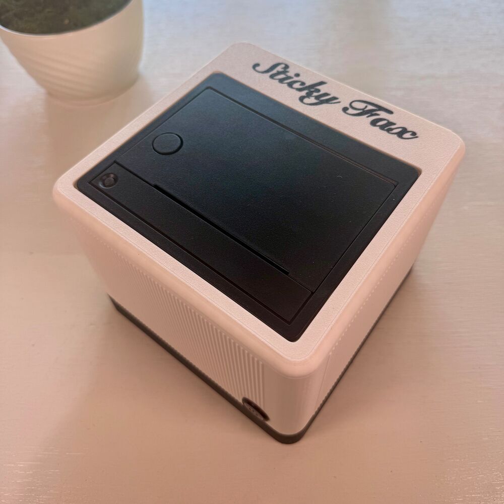 A paper-white StickyFax with an ink-black base and black script logo, on a desk