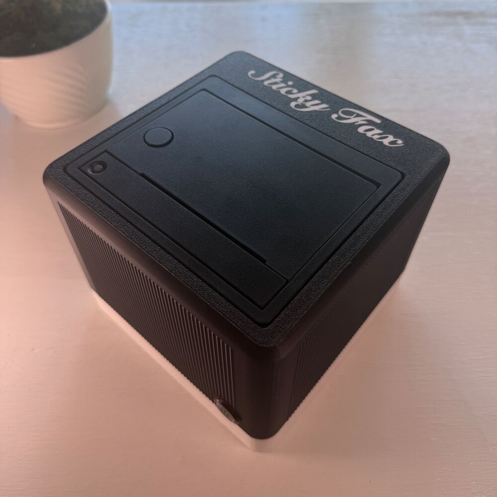 An ink-black StickyFax with a paper-white base and white script logo, on a desk